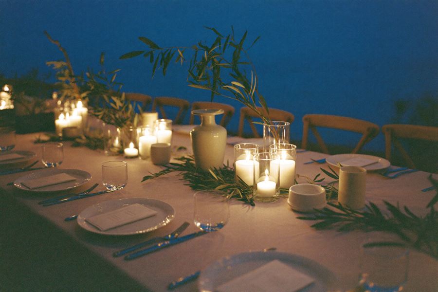 A long linen dinner table set with candles and olive branches at dusk.
