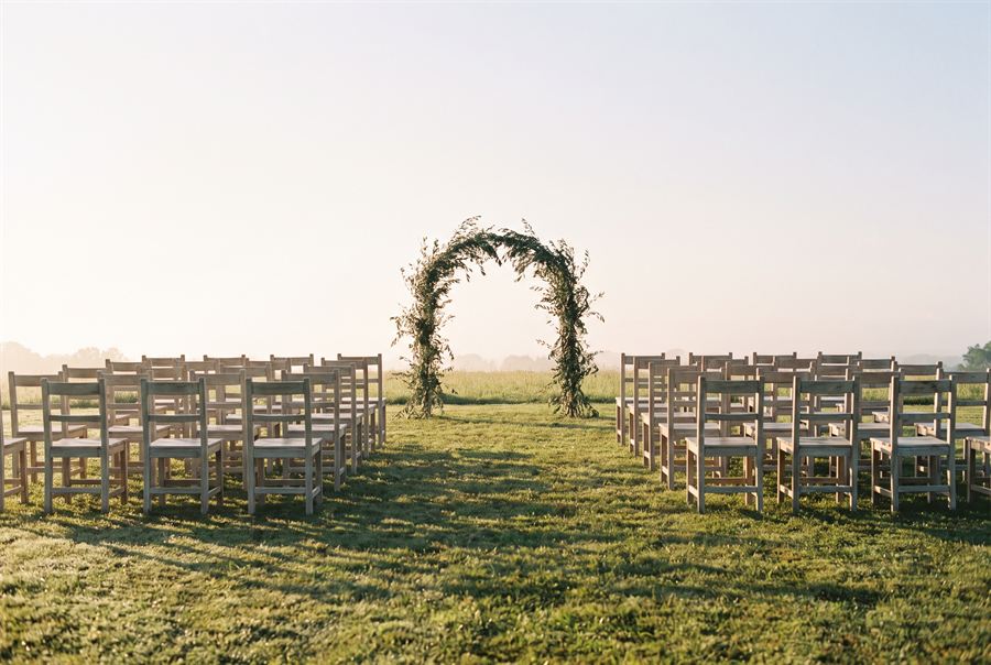 Rows of pale wooden chairs on grass facing an arch of olive branches, before guests arrive.