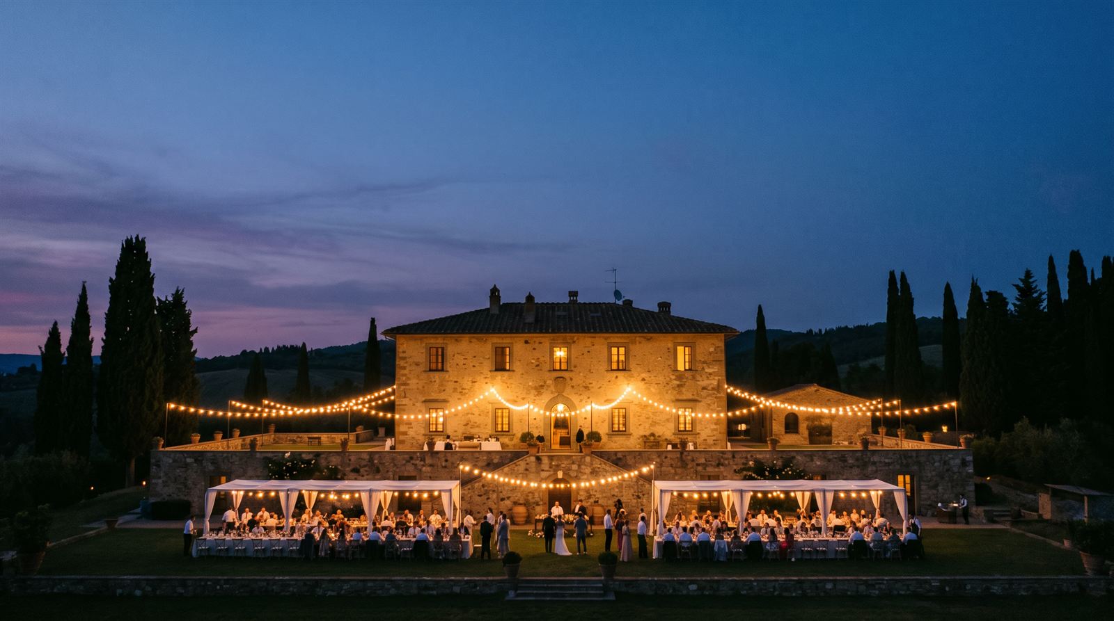 An old stone villa at dusk with warm lit windows and festoon lights strung across a terrace below.